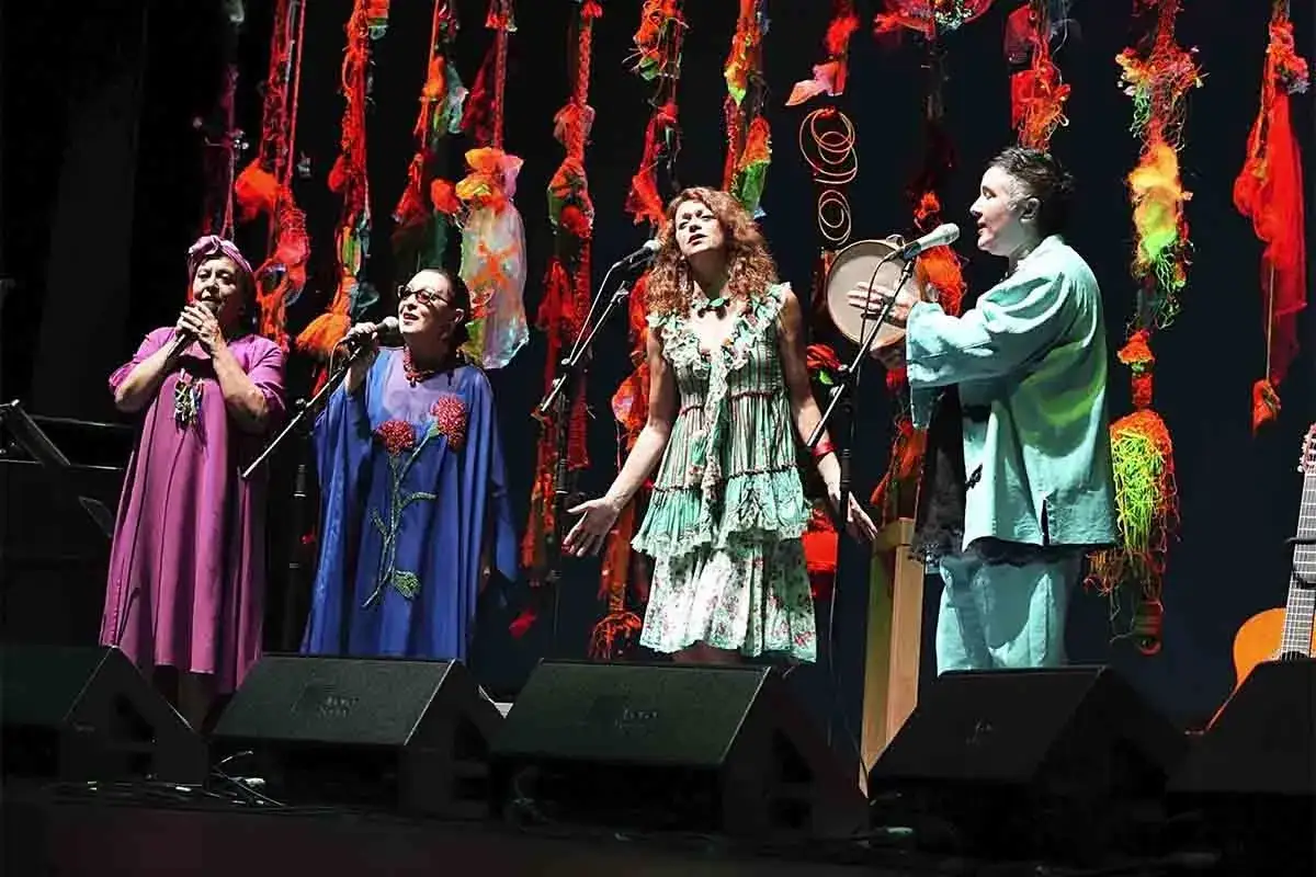 Cuatro mujeres cantan en un escenario con una decoraci&oacute;n vibrante. Una de ellas, con un vestido verde, evoca la jarcha martirio con su expresi&oacute;n.