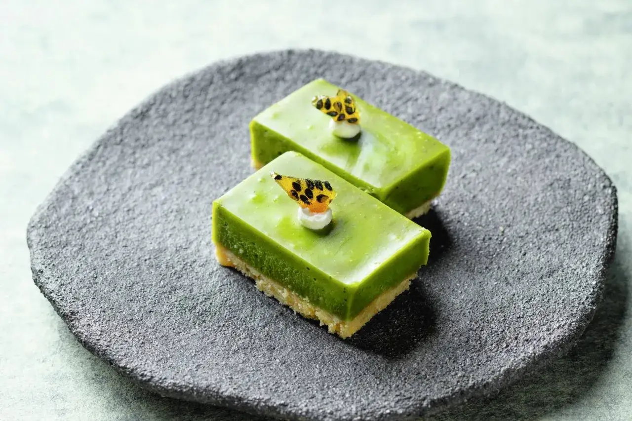 Zwei grüne Matcha Brownies mit einem knusprigen Boden und einem leuchtend grünen Topping, garniert mit einem Tupfer weißer Creme und einem Stück Frucht.