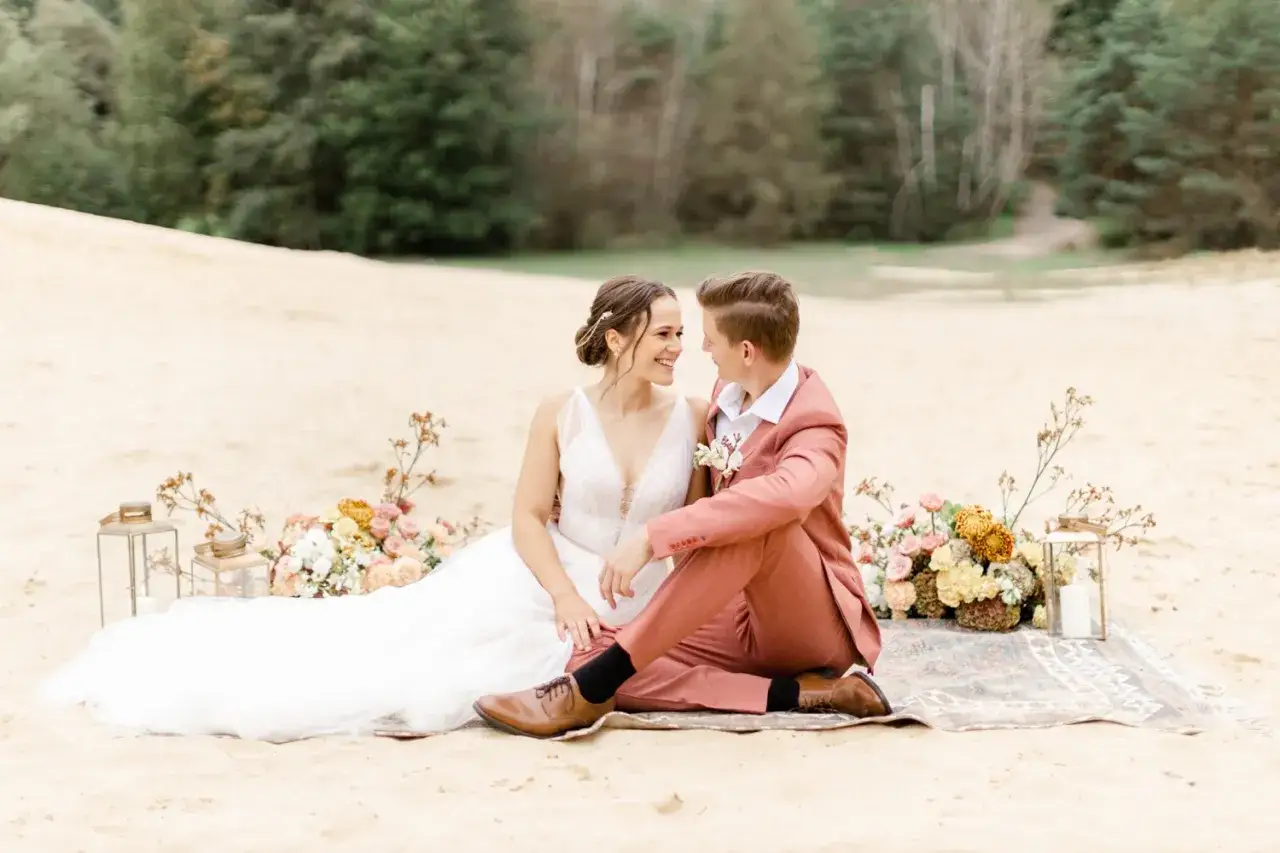 Brautpaar sitzt auf einer Decke im Sand, umgeben von Blumen. Der Br&auml;utigam tr&auml;gt einen korallenfarbenen Anzug. Ein wundersch&ouml;ner Anblick, der den besten Monat zum Heiraten widerspiegelt.