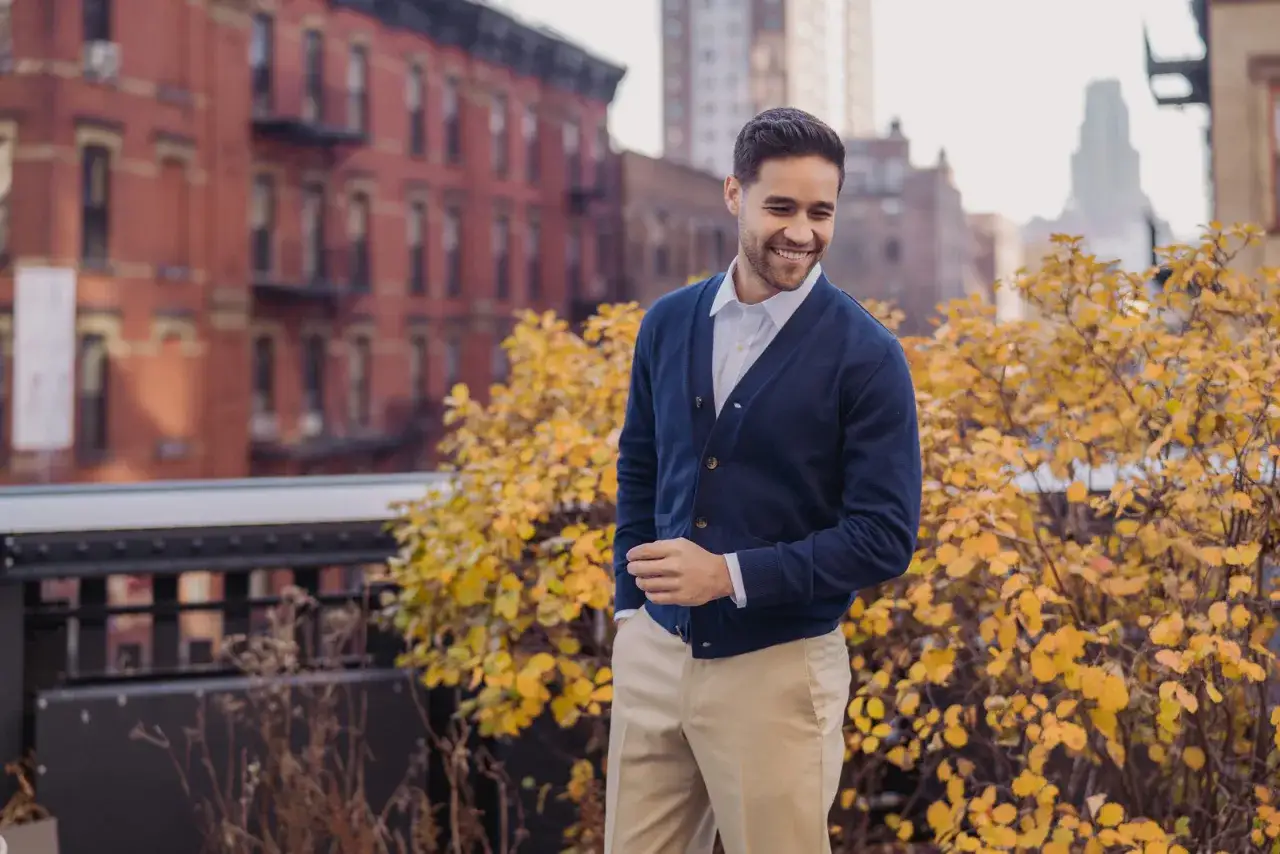 Un homme souriant, en chemise blanche et cardigan bleu marine, montre comment porter un cardigan homme avec élégance, sur fond urbain automnal.