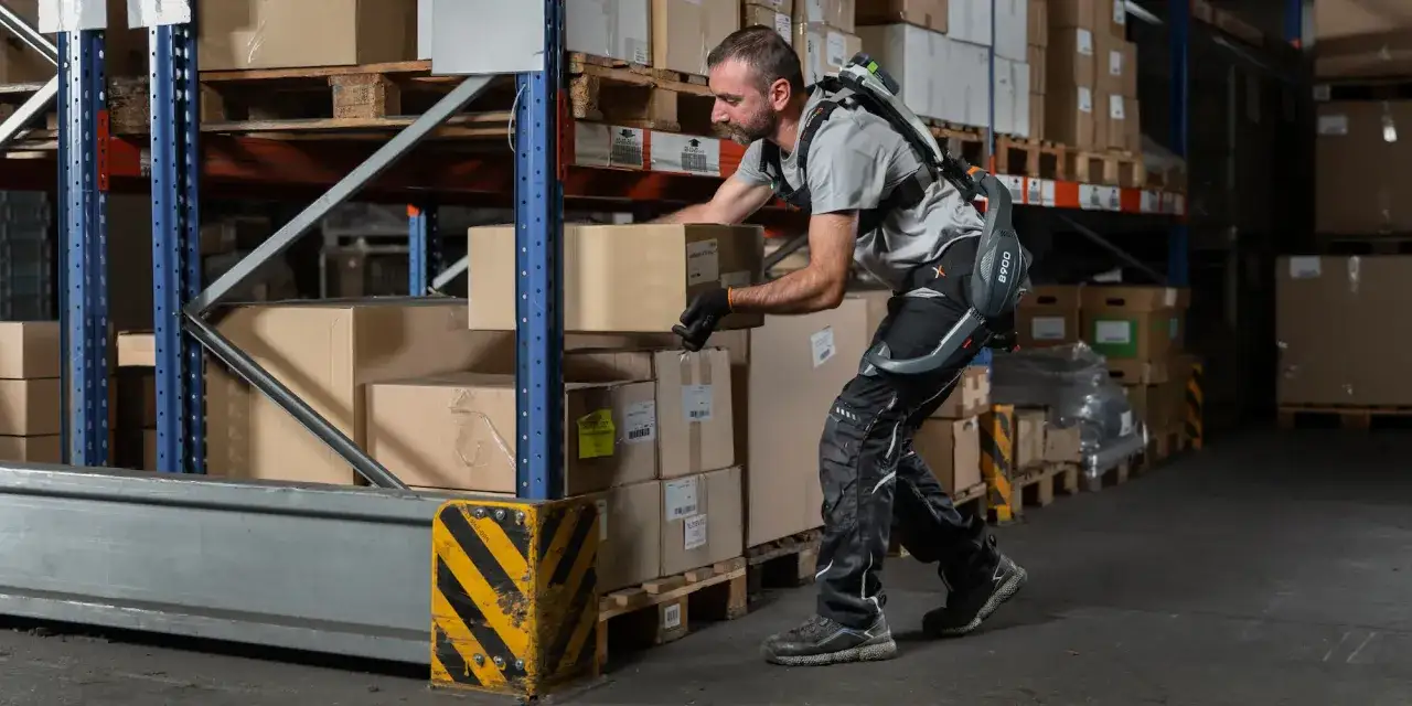 Un homme portant un exosquelette pour aider à la manutention dans un entrepôt. Cet exosquelette est conçu pour réduire la charge sur le corps, offrant un soutien aux personnes ayant un handicap ou effectuant des tâches physiques lourdes.