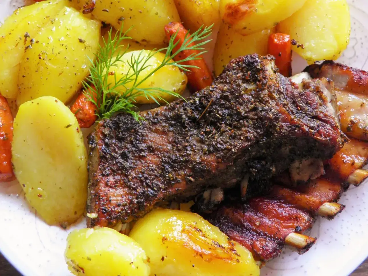 Pieczona żeberka z ziemniakami i marchewką, udekorowane koperkiem. Idealne danie obiadowe, które świetnie smakuje z tymi dodatkami.