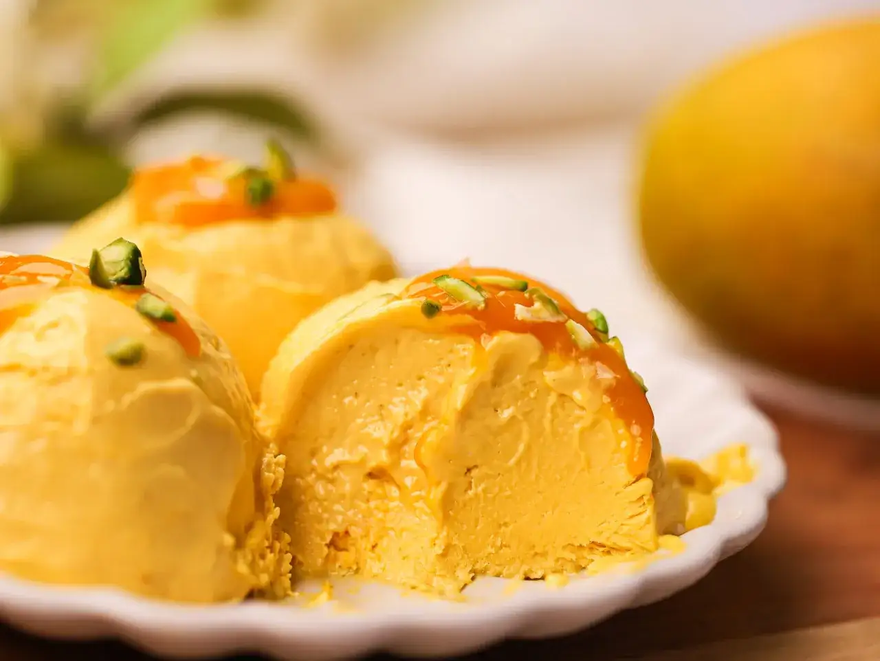 Cremiges Mango Eis mit Orangensauce und gehackten Pistazien auf einem weißen Teller. Ein ganzer Mango liegt im Hintergrund.