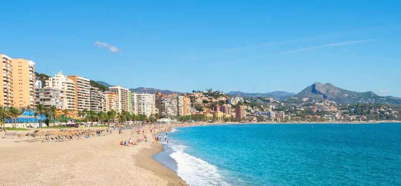 Sonnige Str&auml;nde und hohe Geb&auml;ude s&auml;umen die K&uuml;ste der Costa del Sol. Menschen entspannen sich am Sandstrand, w&auml;hrend das t&uuml;rkisfarbene Meer sanft anrollt.