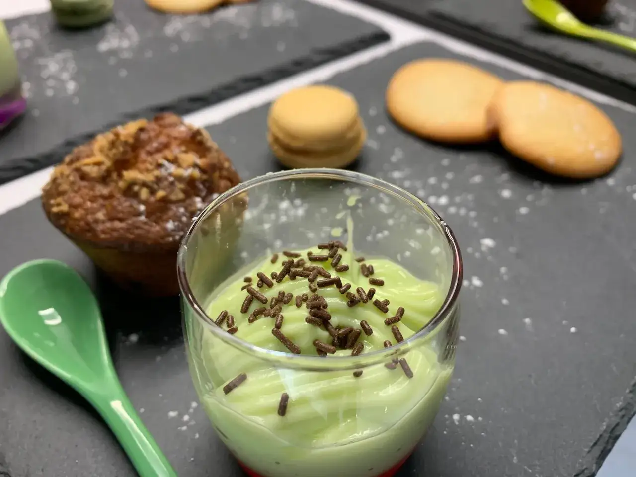 Douceur verte : une crème anglaise pistache onctueuse, saupoudrée de vermicelles chocolat, à côté d'un muffin et de macarons.