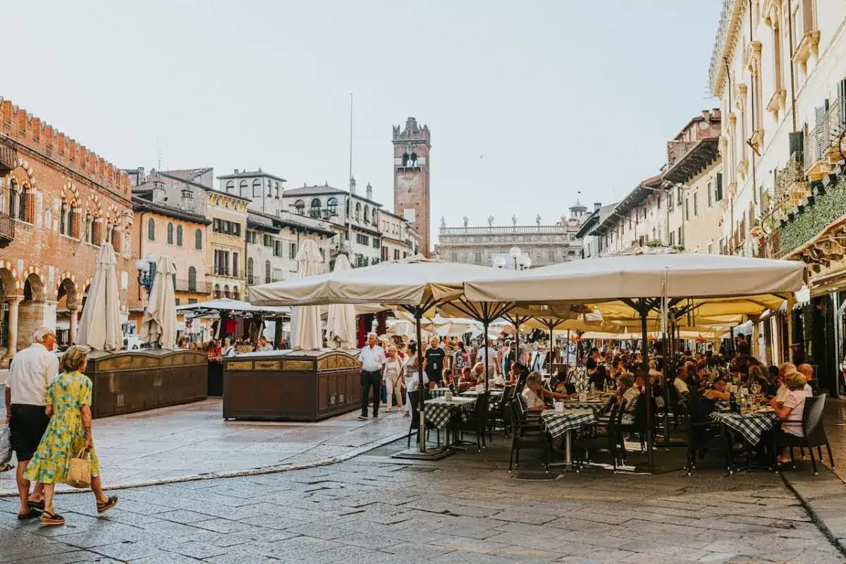 Piazza affollata a Verona, con tavoli all'aperto per un piacevole aperitivo. Persone passeggiano tra gli edifici storici.