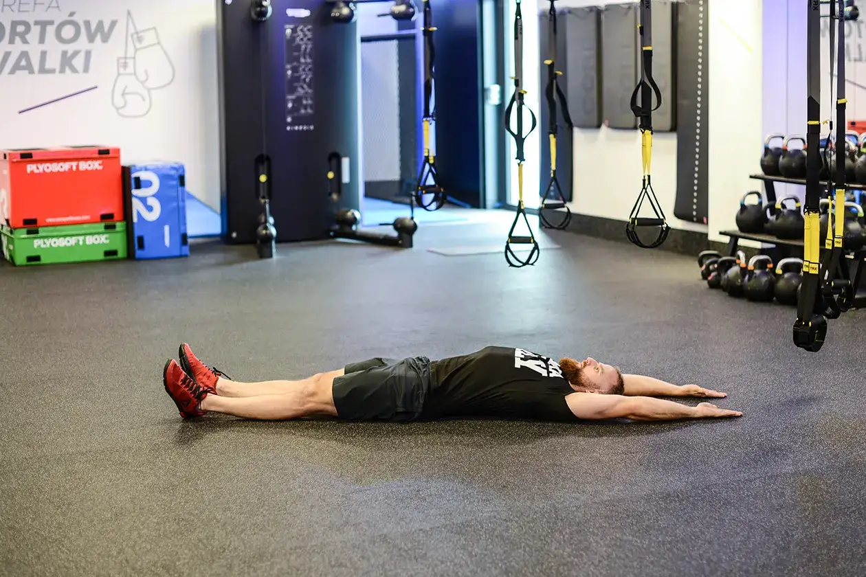 Mężczyzna wykonuje ćwiczenie na brzuch, leżąc na macie z wyciągniętymi rękami. W tle widać sprzęt treningowy, w tym skrzynie plyo i kettlebells.