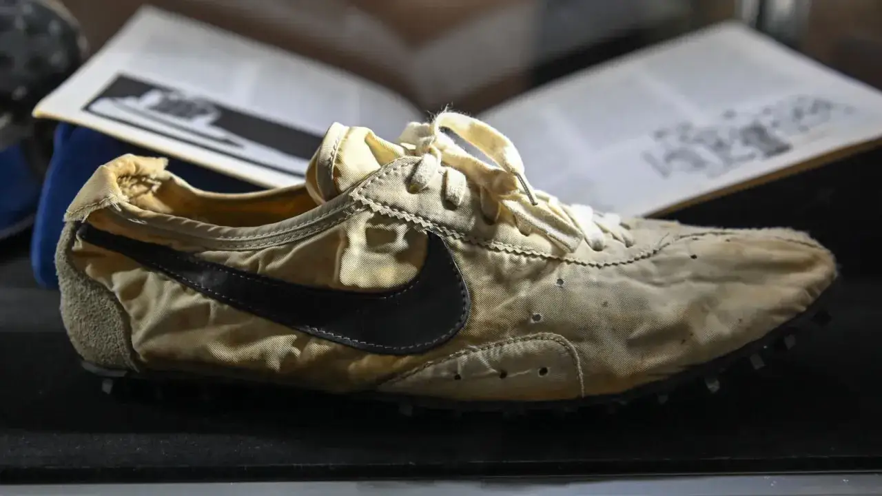 Stary, beżowy but Nike z czarnym logo, prawdopodobnie najdroższe buty Nike w historii, leży obok otwartej księgi.