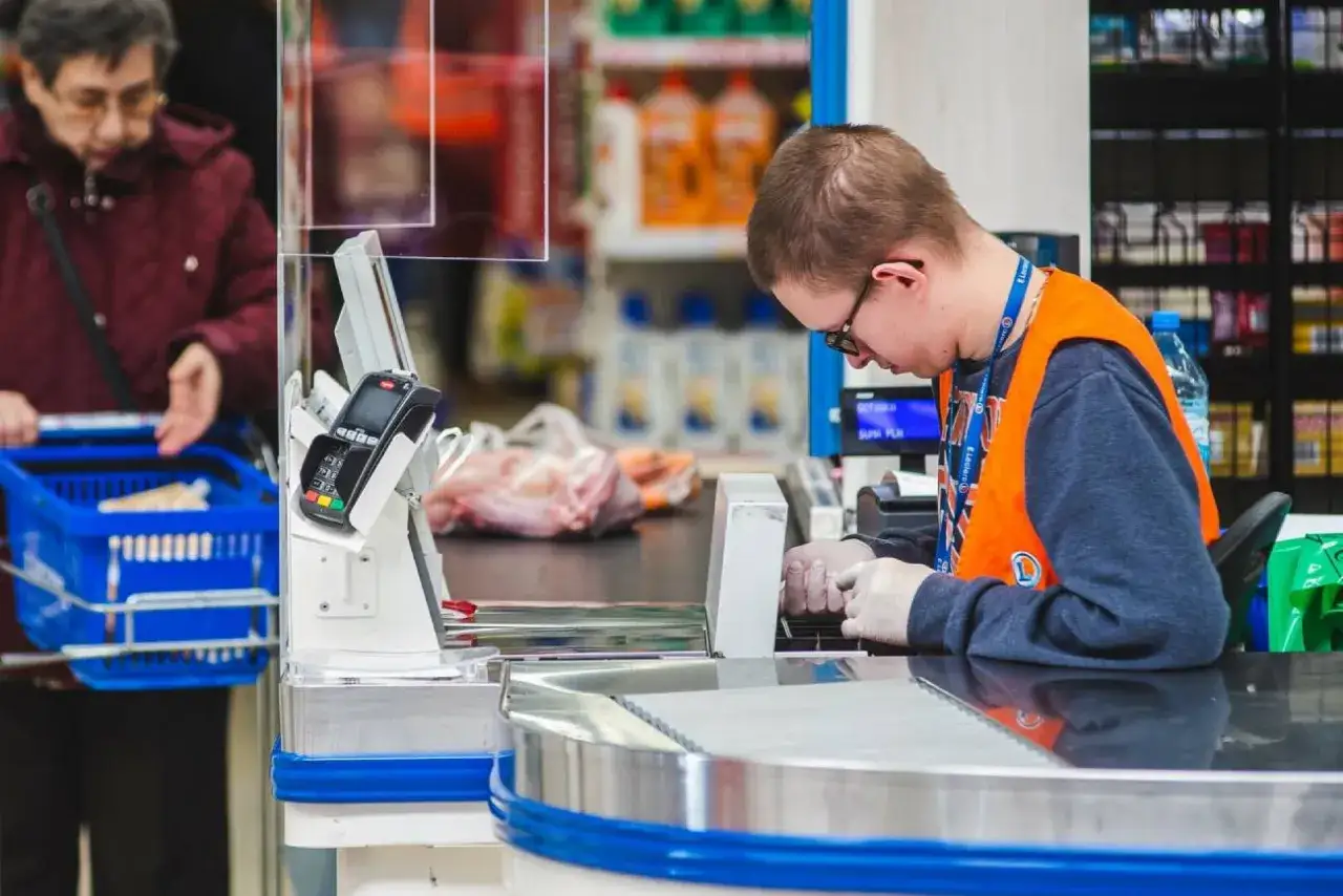 Kasjer w Żabce w pomarańczowej kamizelce obsługuje klienta. Ile zarabia pracownik Żabki netto? To pytanie nurtuje wielu.