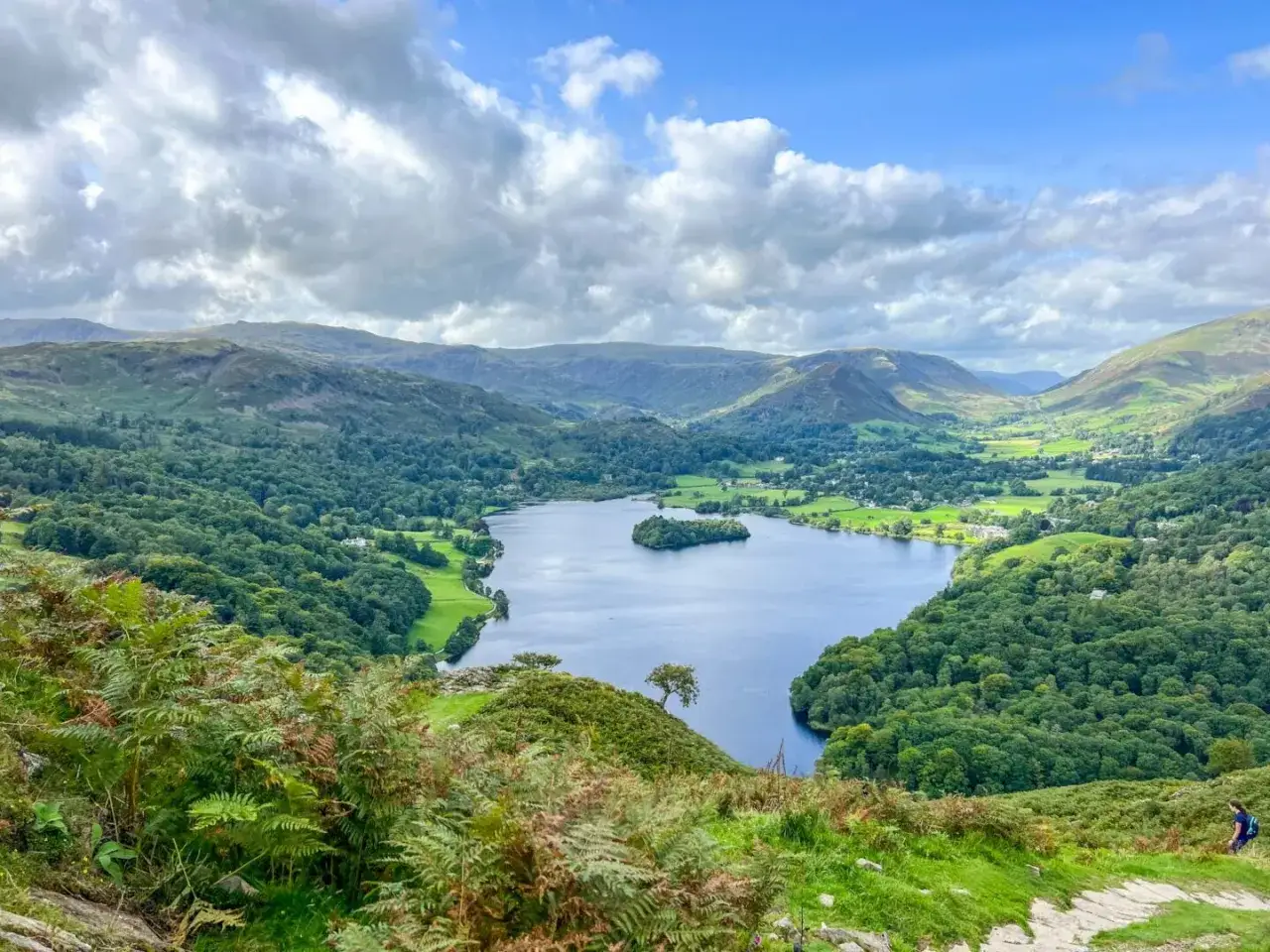 A stunning view of a lake surrounded by green hills and forests, perfect for day trips from Liverpool. A lone hiker walks on a path in the foreground.