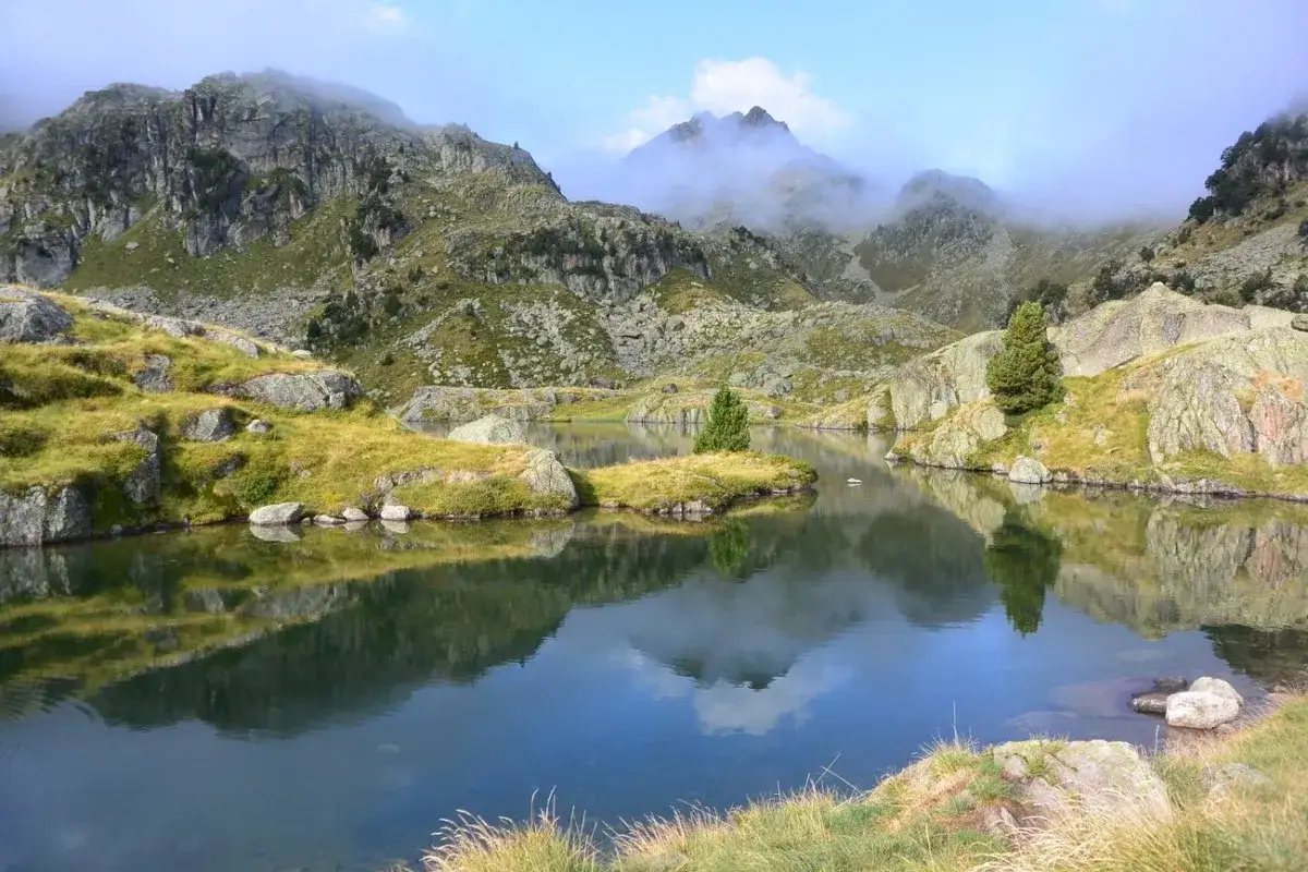 Calme lac reflétant les montagnes rocheuses et verdoyantes du cirque de Colomers, avec un ciel bleu et des nuages bas.