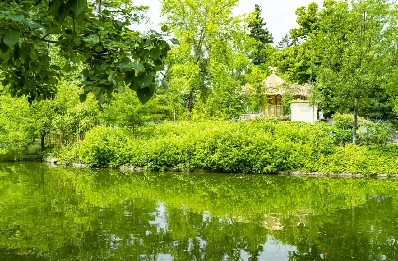 Un carrousel doré scintille au milieu d'une végétation luxuriante, reflété dans l'eau paisible d'un parc floral à Bordeaux.