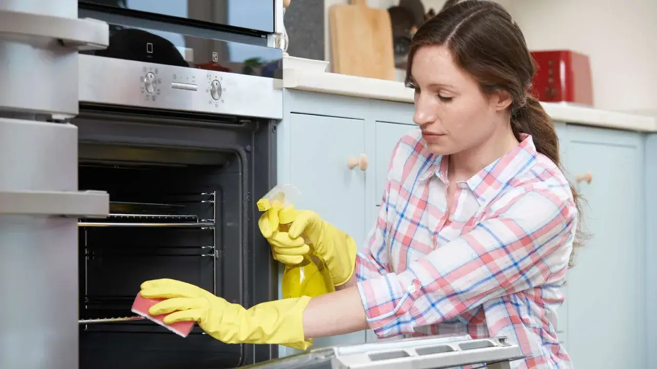 Frau mit gelben Handschuhen reinigt Backofen mit Spr&uuml;hflasche und Schwamm. Sie macht Backofenreiniger selber.