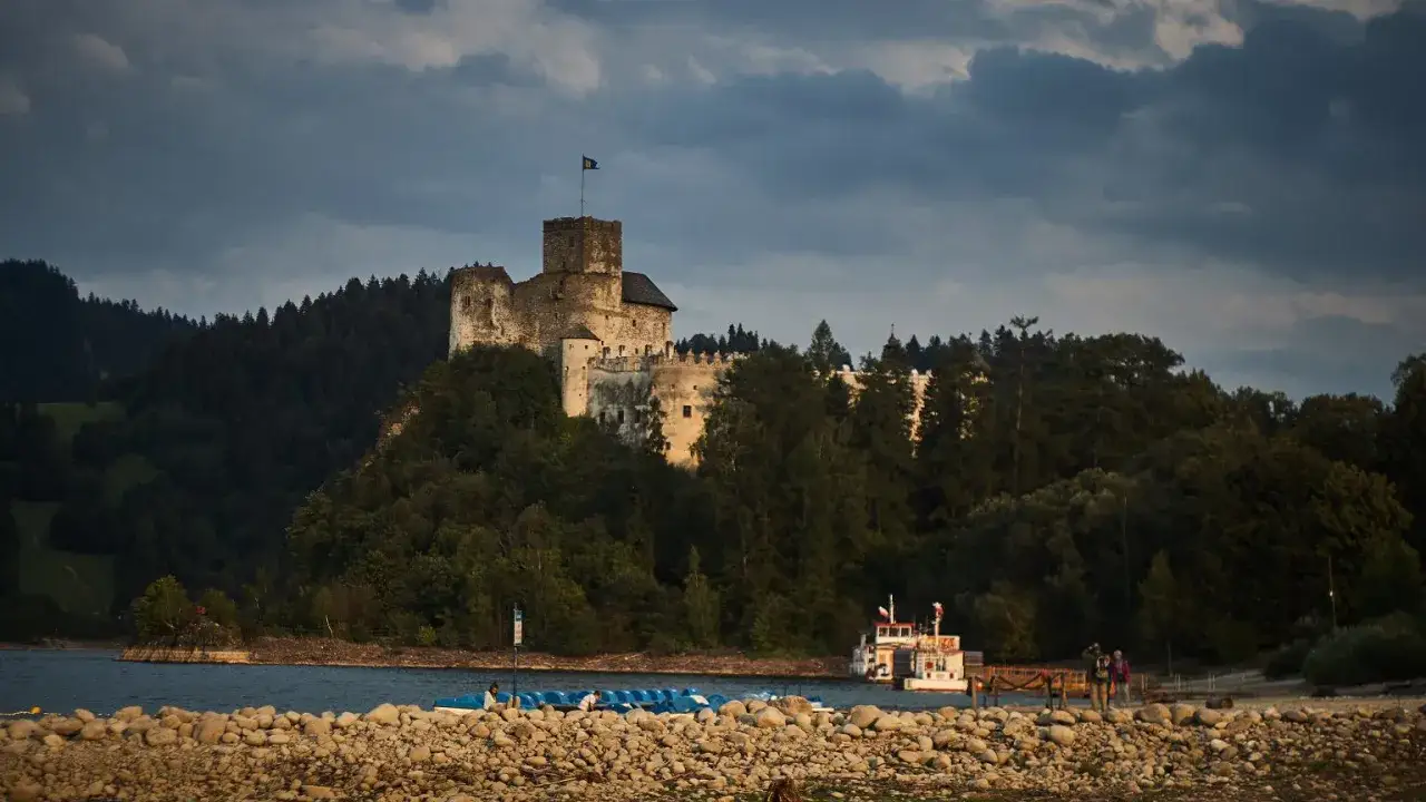 Zamek na wzgórzu nad jeziorem, otoczony lasem. W oddali widać statek wycieczkowy. To malowniczy krajobraz w okolicach najstarszego parku narodowego w Polsce.