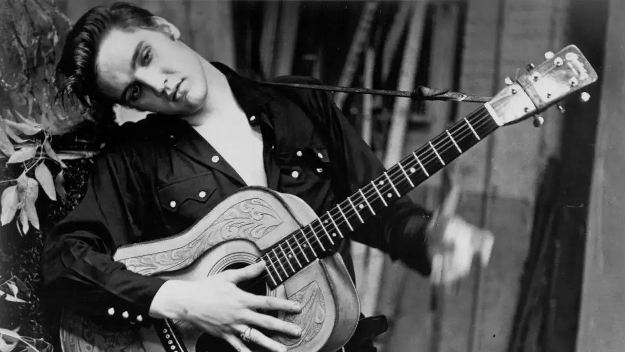 Junger Elvis Presley mit Gitarre, sein Kopf ruht auf seinem Arm, w&auml;hrend er sich an einen Baum lehnt.