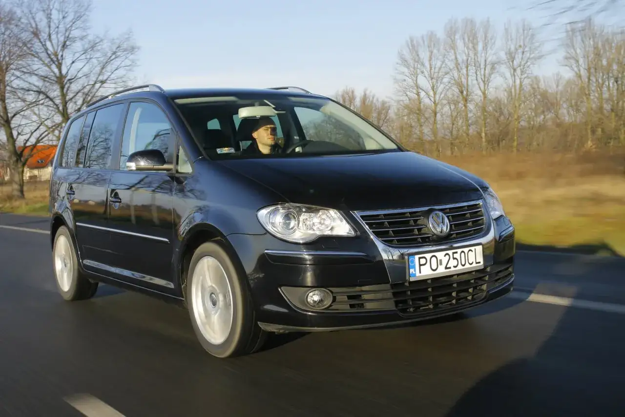 Czarny VW Touran jedzie drogą, widać jego przednie światła i grill.