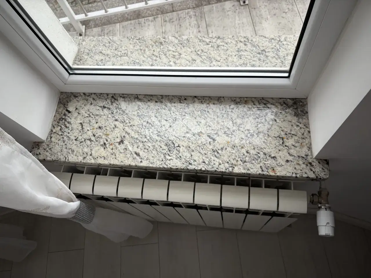 Elegancki parapet z konglomeratu granitowego nad białym grzejnikiem, ozdobiony delikatną firanką.