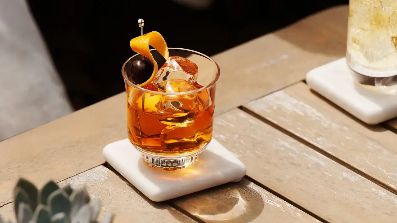 Ein Glas mit bernsteinfarbenem Whiskey neat, Eisw&uuml;rfeln, einer Kirsche und Orangenzeste auf einem Holztisch.