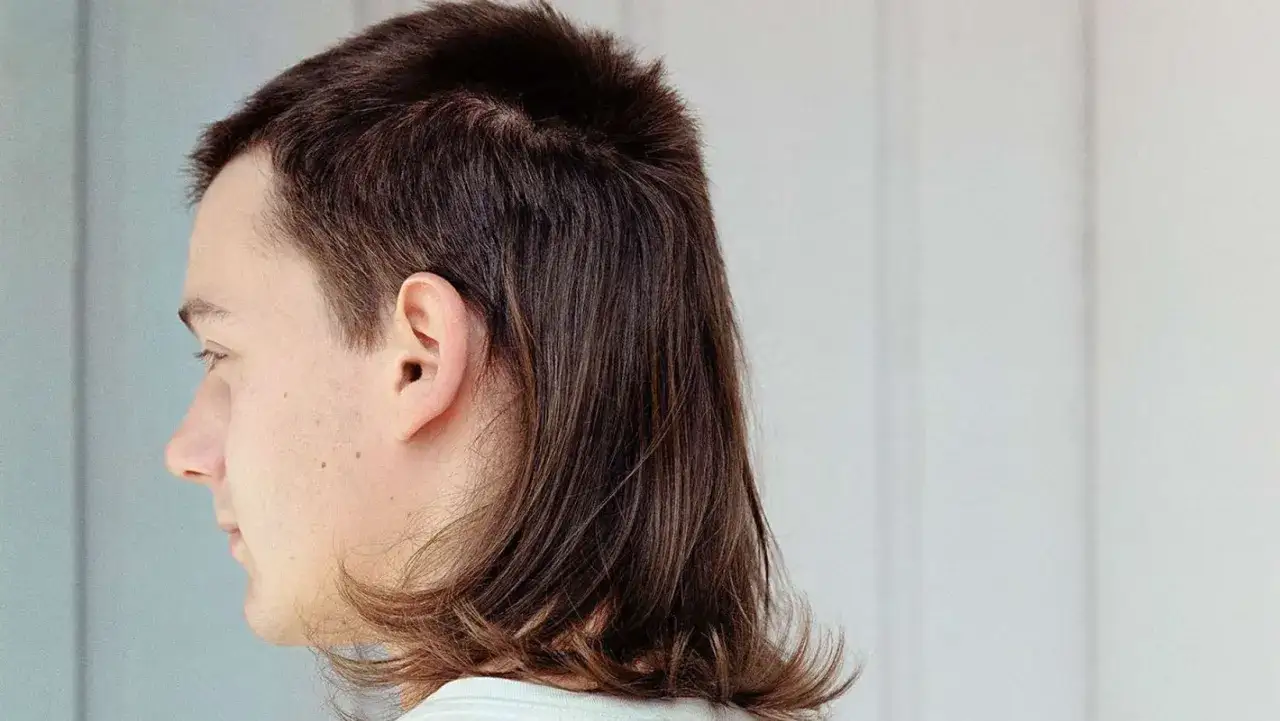 Młody mężczyzna z charakterystyczną fryzurą mullet. Krótsze włosy z przodu i dłuższe z tyłu nadają mu stylowy wygląd.