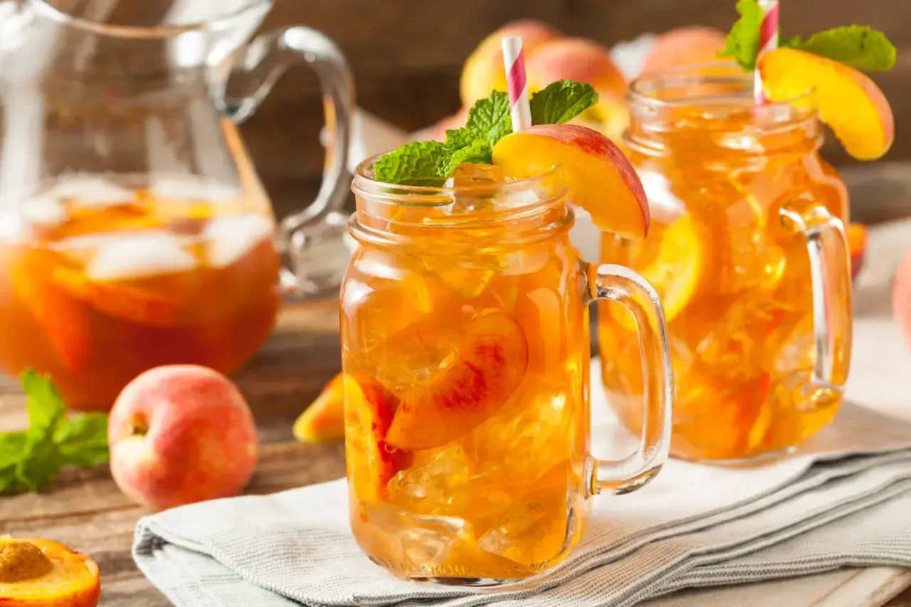 Erfrischender Pfirsich Eistee selber machen: Gläser mit Eis, Pfirsichscheiben und Minze, daneben eine Karaffe mit dem leuchtend orangen Getränk.