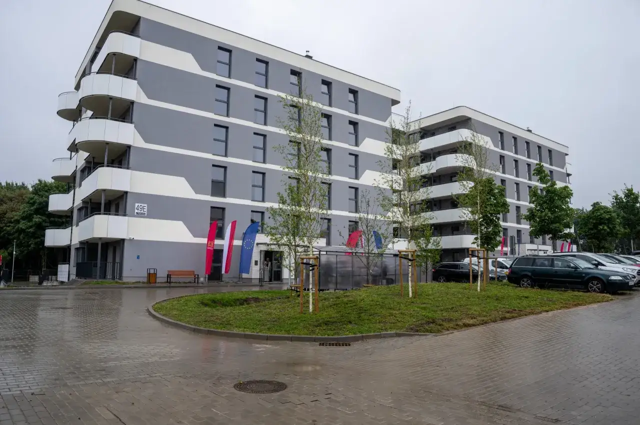 Nowoczesne mieszkanie TBS z balkonami, szaro-białą elewacją, flagami i parkingiem.