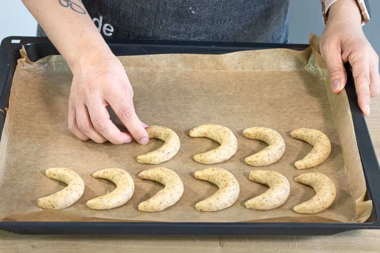 Hände legen frisch geformte Vanillekipferl auf ein Backblech. Die Halbmonde duften schon nach Vanille und Mandeln.