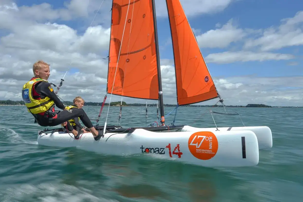 Deux jeunes garçons naviguent sur un petit voilier pour débutant, leurs voiles oranges gonflées par le vent sous un ciel bleu et nuageux.