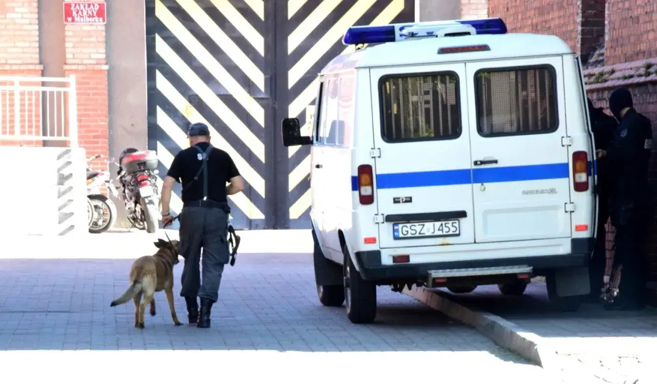 Funkcjonariusz z psem policyjnym idzie w kierunku więzienia. Nie wiadomo, po jakim czasie policja doprowadza do zk.