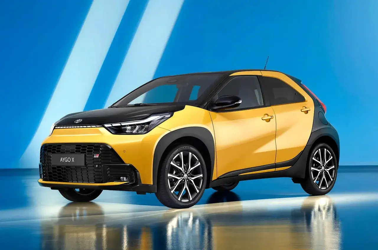 Żółty Toyota Aygo X Hybrid z czarnym dachem i maską, prezentujący się dynamicznie na tle niebieskich pasów.