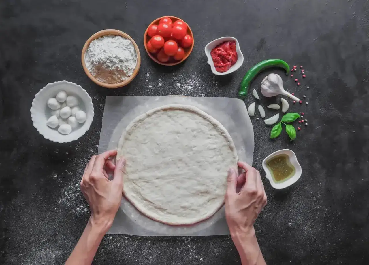 Ręce formują okrągłe ciasto na pizzę. Obok mąka, pomidorki, mozzarella, sos pomidorowy, czosnek, papryczka i bazylia.