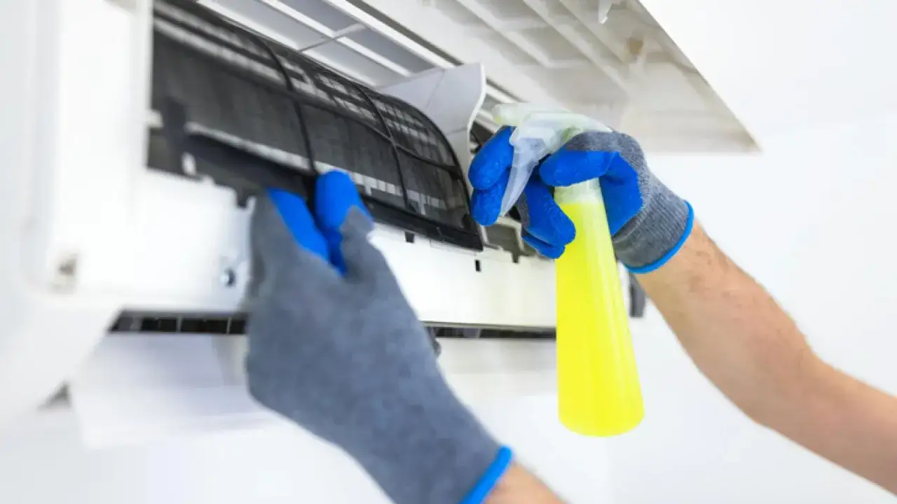 Manos enguantadas sacando el filtro de un aire acondicionado para limpiarlo con spray.