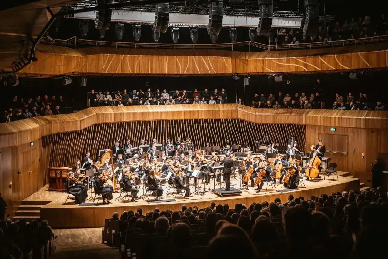 Orkiestra symfoniczna gra uwertura w nowoczesnej sali koncertowej. Widownia w skupieniu słucha muzyki.