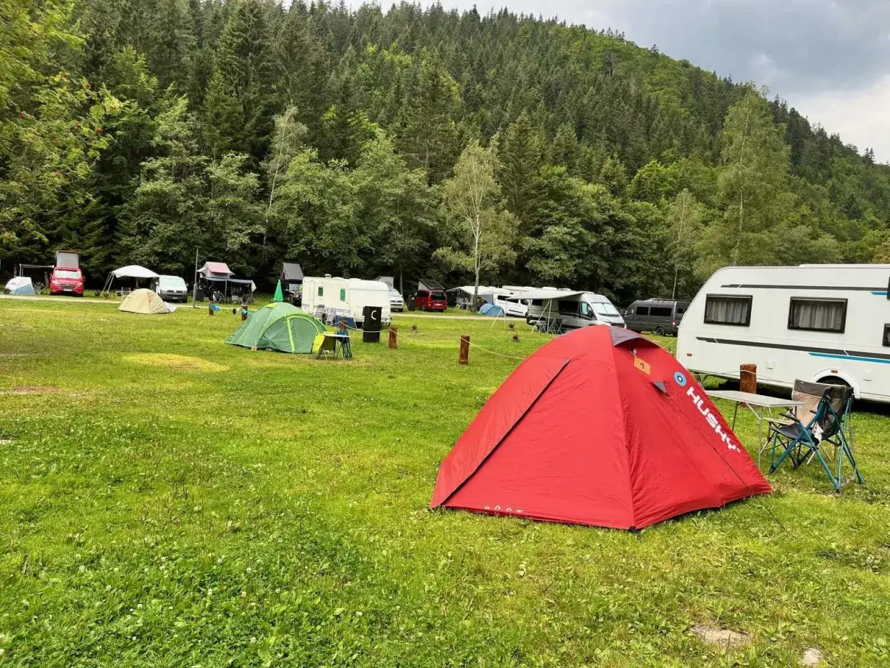 Czeska Szwajcaria camping: czerwony namiot Husky na trawie, zielony namiot, kampery i las w tle.