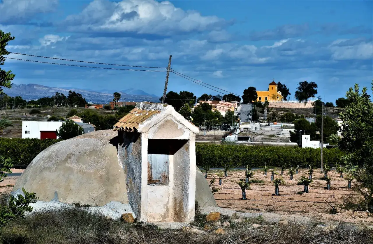 Antiguo aljibe de agua con una peque&ntilde;a caseta, rodeado de vegetaci&oacute;n y un campo de frutales. Al fondo, un pueblo con una iglesia amarilla.