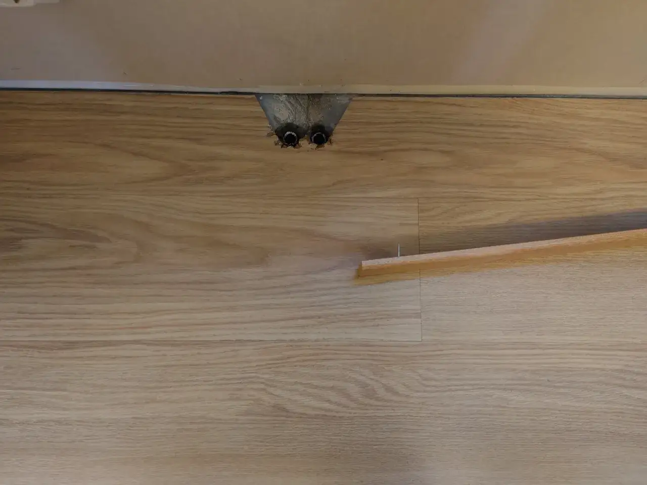 Här syns en del av ett laminatgolv med synliga rörgenomföringar. En list och en spik ligger redo för att laga laminatgolv.