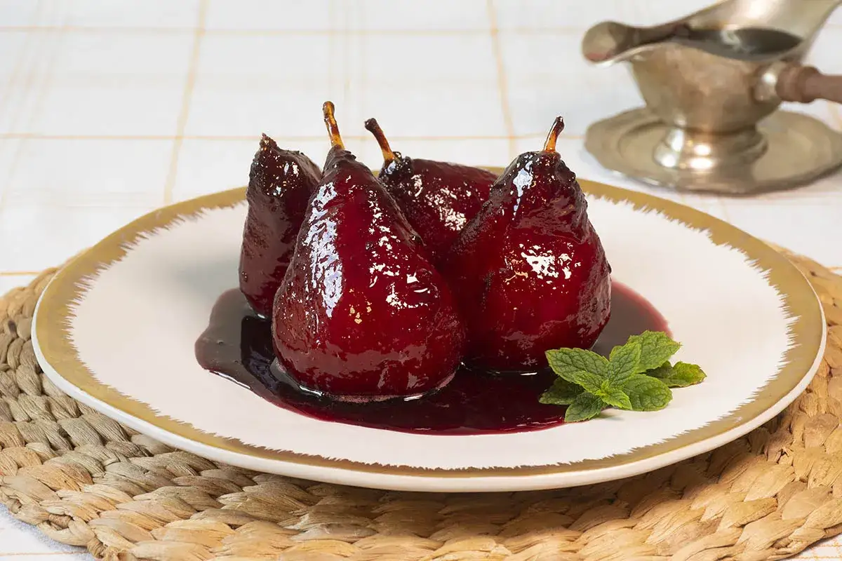 Cuatro peras al vino, brillantes y oscuras, servidas en un plato blanco con borde dorado, bañadas en su propio almíbar.
