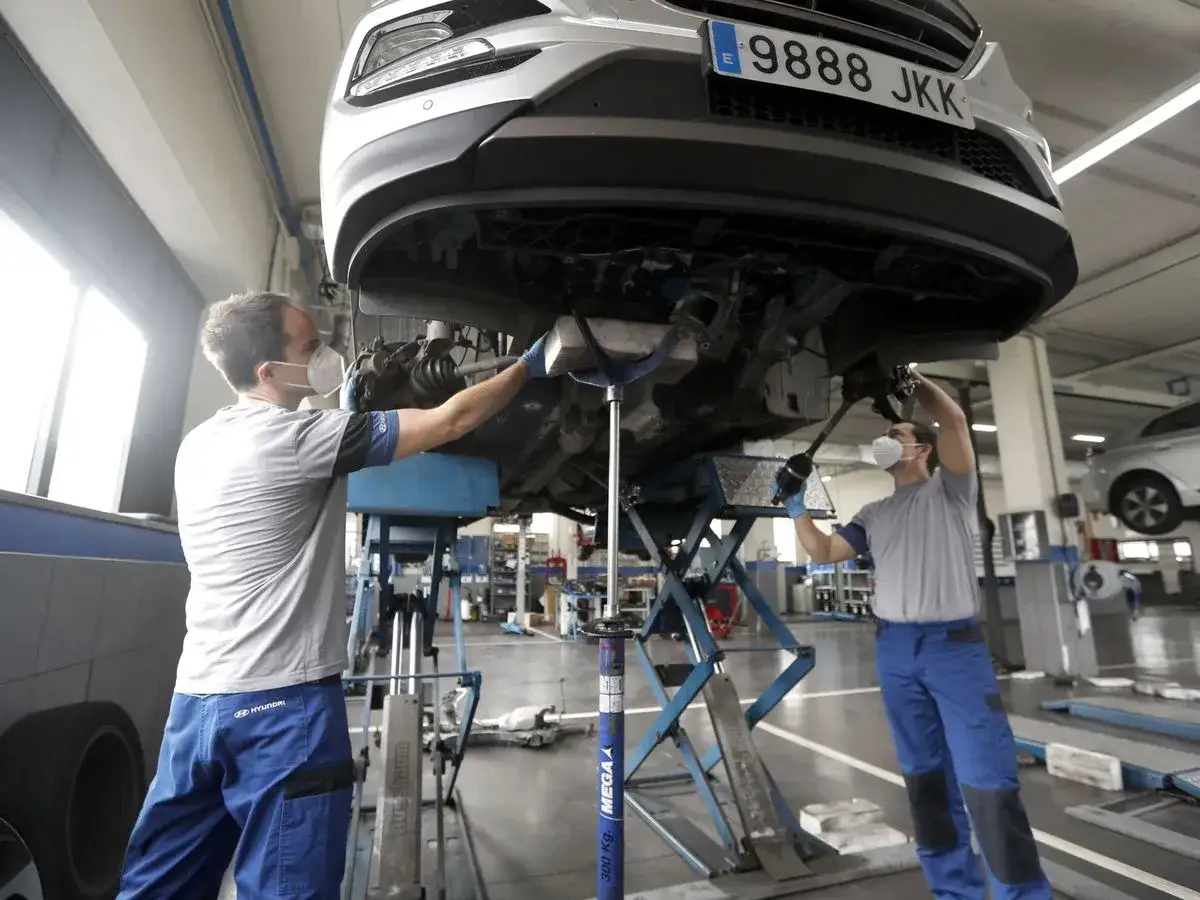Dos mecánicos con mascarilla trabajan en el tren delantero de un coche elevado. Consulta el precio del servicio de mantenimiento del coche.