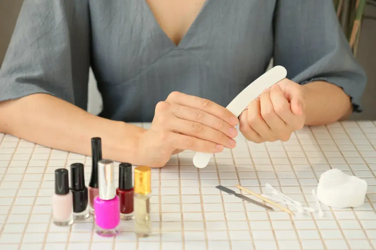 Kobieta wykonuje **manicure klasyczny**, opiłowując paznokcie. Na stole stoją kolorowe lakiery i akcesoria do pielęgnacji.