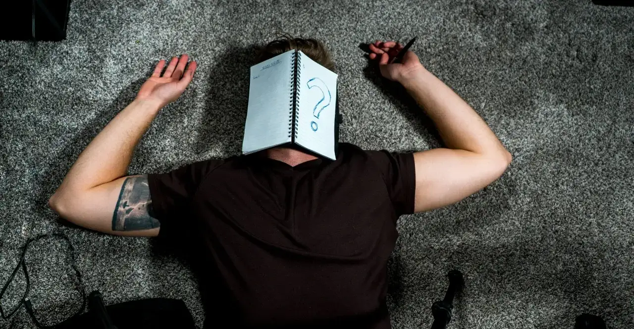 Persona tirada en el suelo, con un cuaderno abierto sobre la cara que muestra un gran signo de interrogaci&oacute;n. Parece que busca temas para hacer una redacci&oacute;n.