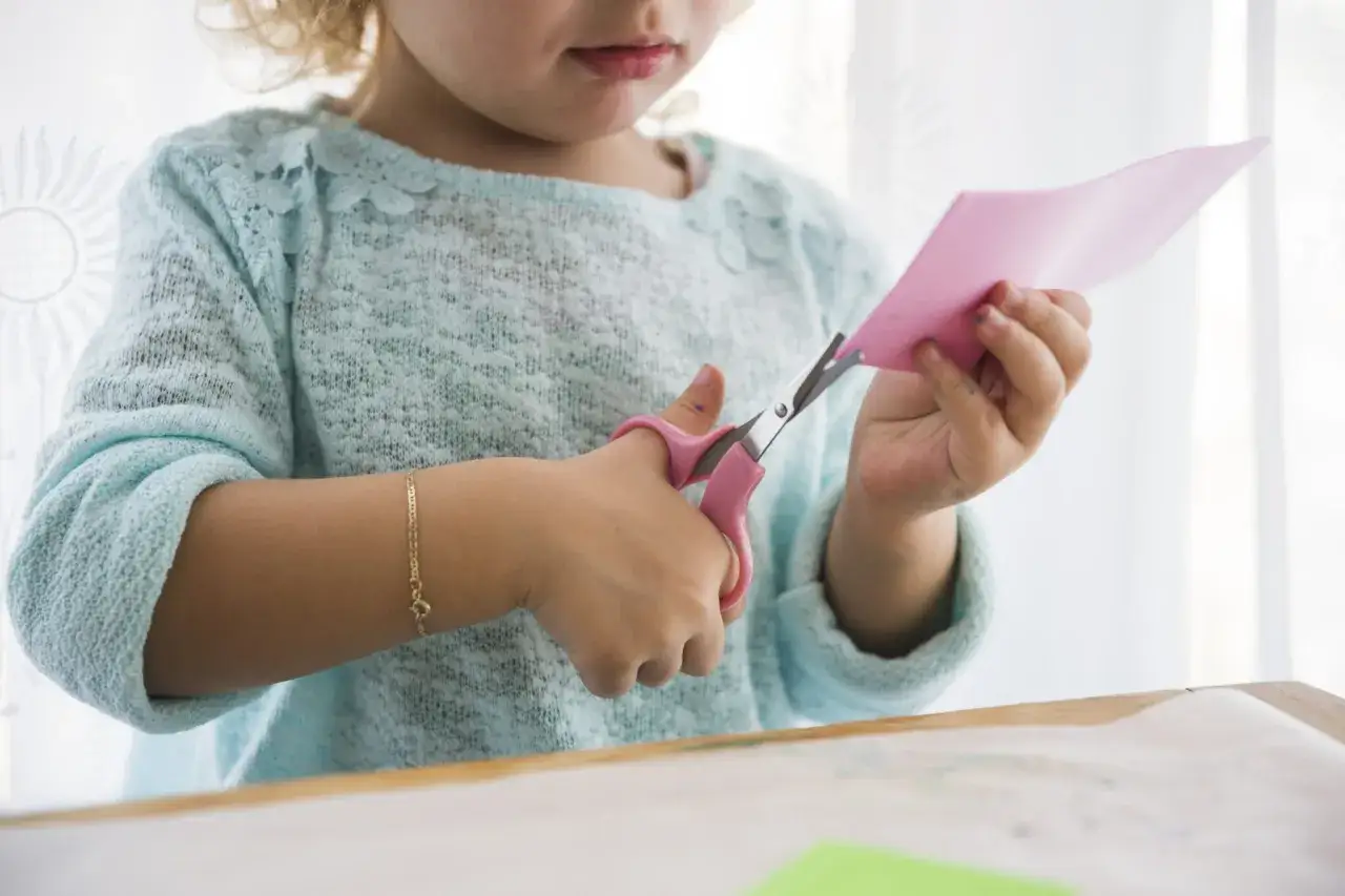 Mała dziewczynka ćwiczy naukę wycinania karty pracy, trzymając r&oacute;żowe nożyczki i r&oacute;żowy papier.