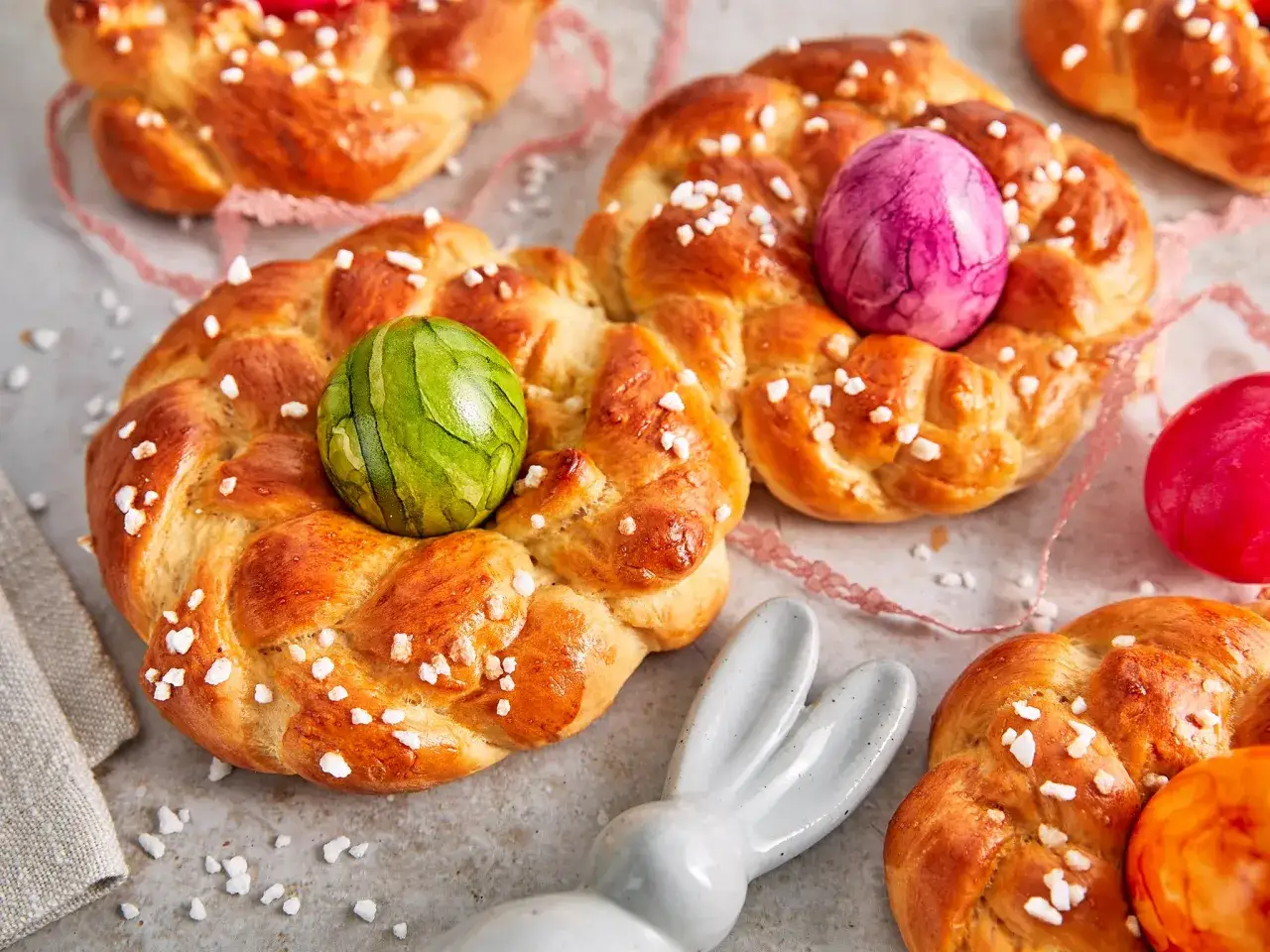 Goldbraune Hefezöpfe mit Zuckerkristallen und bunten Ostereiern, perfekt zum Osterfest. Das Backen dieser Osternester ist eine schöne Tradition.
