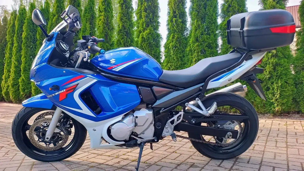 Niebieski motocykl Suzuki GSX650F z czarnym kufrem na bagaż, zaparkowany na bruku przed żywopłotem.