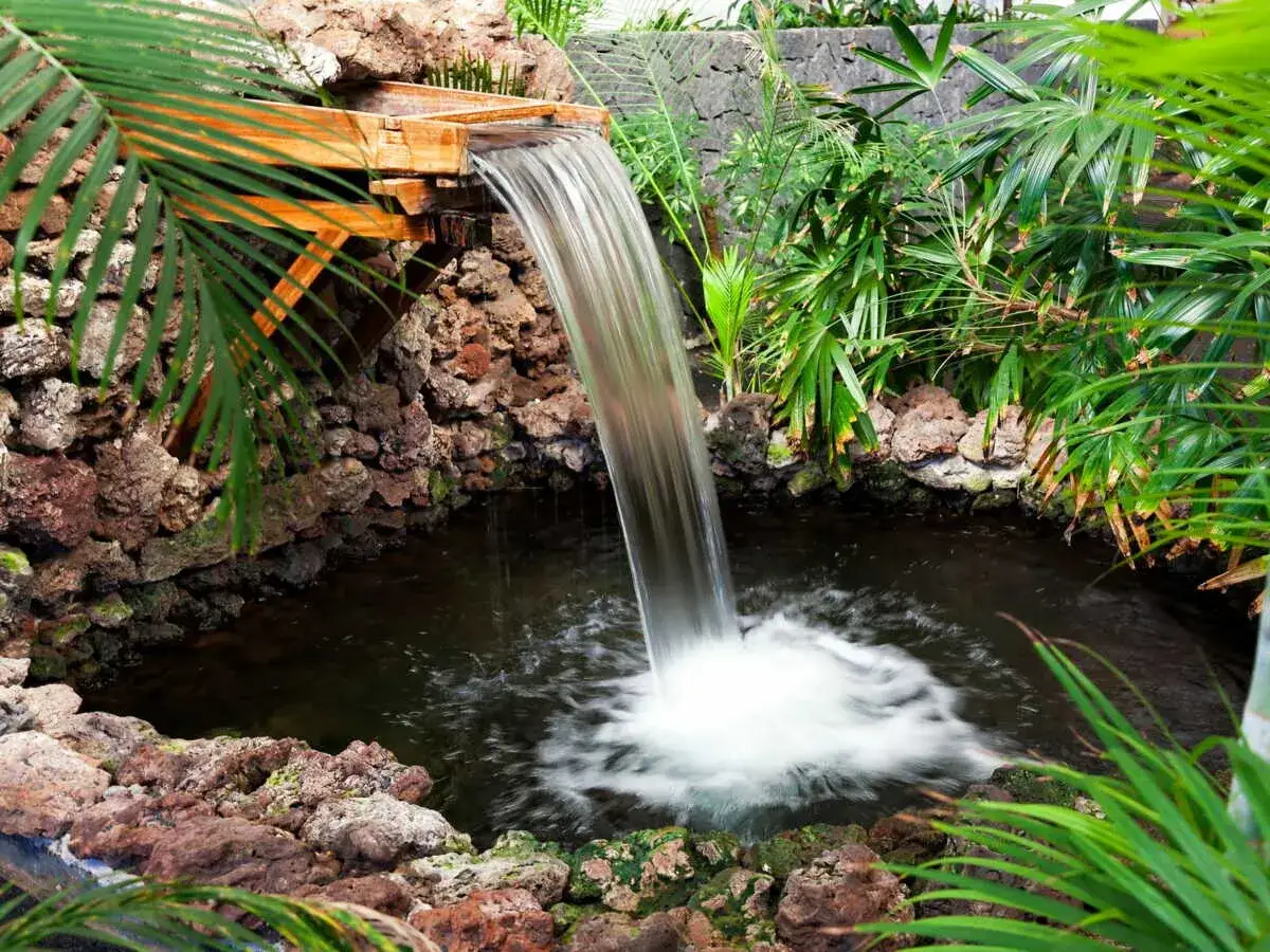 Une cascade en bois, entour&eacute;e de roches volcaniques et de v&eacute;g&eacute;tation luxuriante, tombe dans un petit bassin.