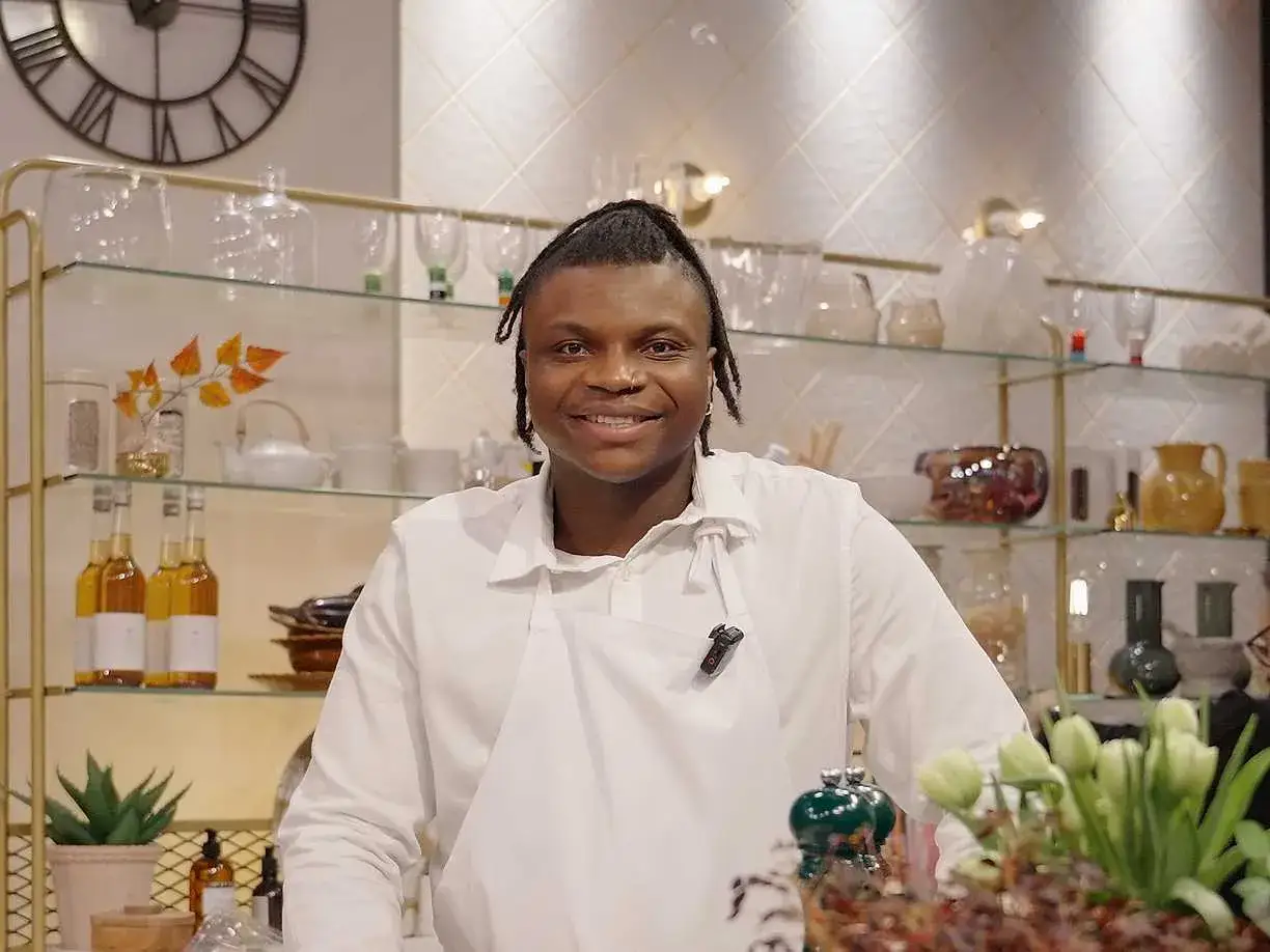 Kelvin Wokomaty, en glad kock med flätor, ler bakom en disk fylld med blommor och flaskor.
