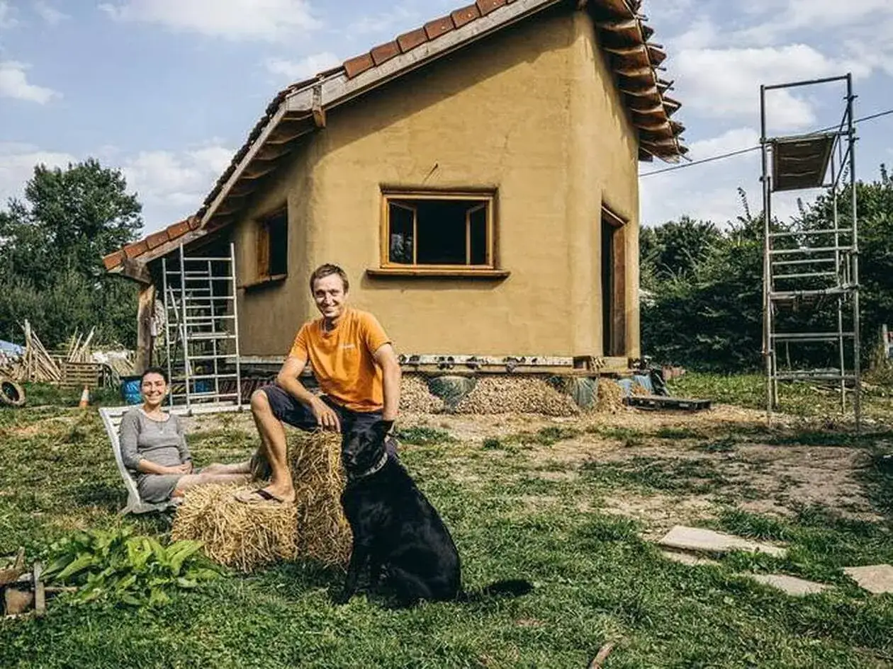 Un couple et leur chien profitent du soleil devant leur maison en **mur terre paille**. L'architecture &eacute;cologique est en cours de finition.