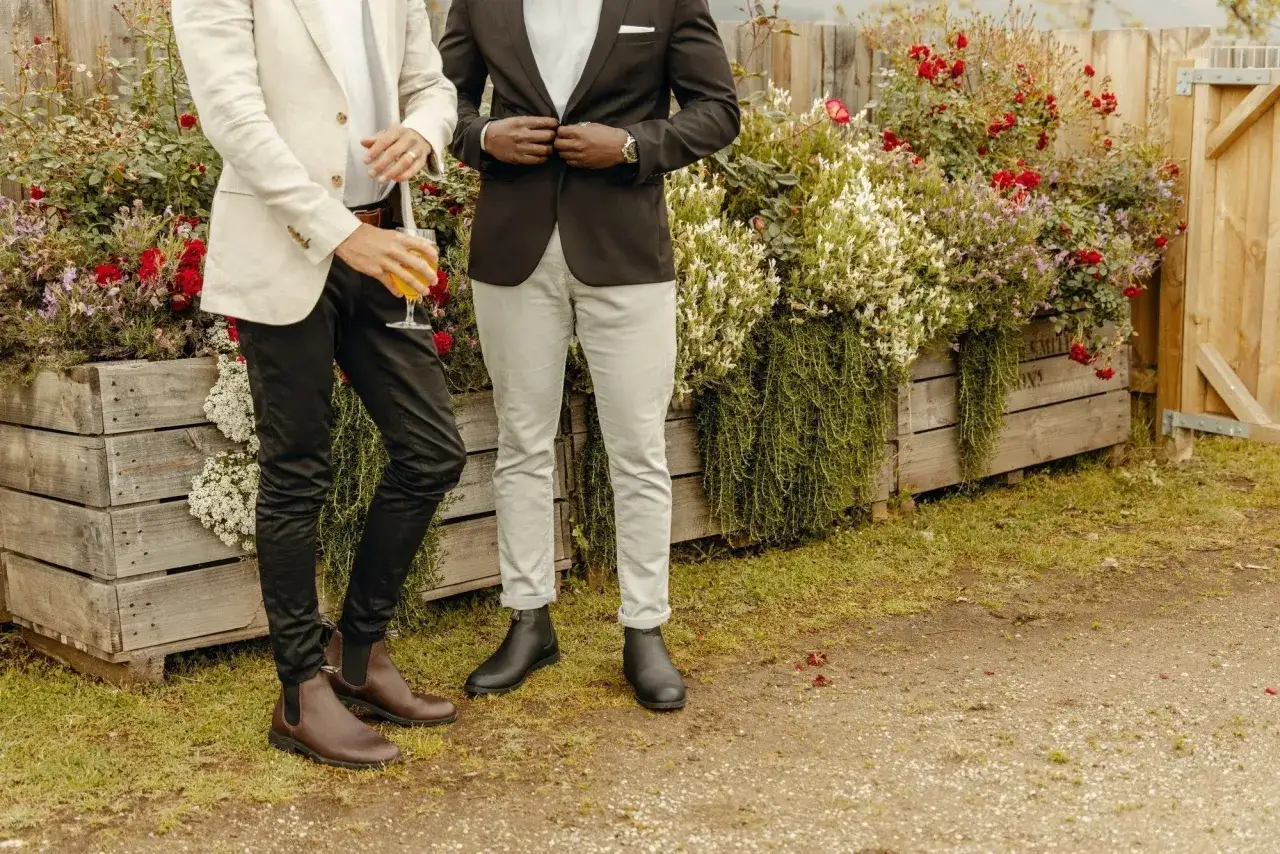 Zwei M&auml;nner in Anz&uuml;gen, einer mit braunen Chelsea Boots, der andere mit schwarzen, stehen vor bl&uuml;henden Holzkisten.