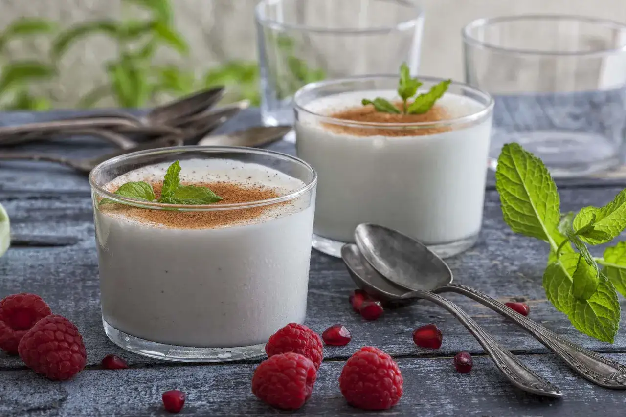 Zwei Gläser cremiger Leche Merengada, garniert mit Minze und Zimt, umgeben von Himbeeren und Granatapfelkernen.