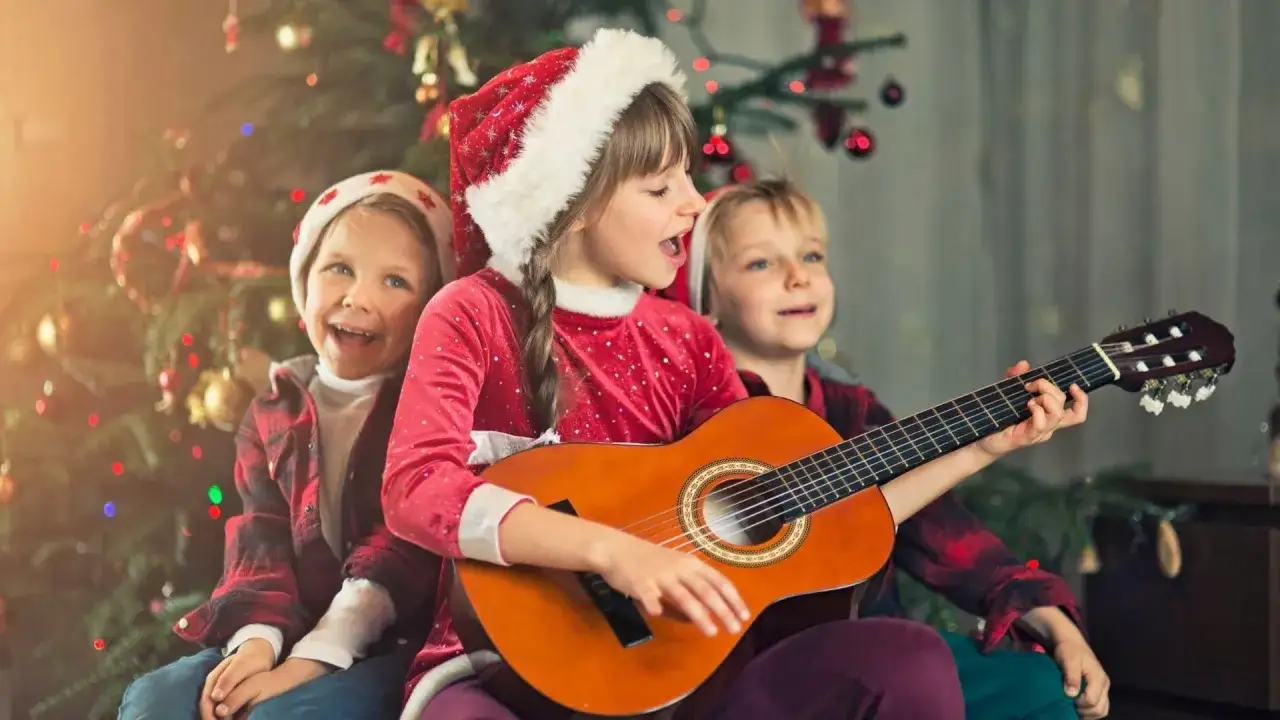 Dzieci w świątecznych czapkach śpiewają kolędy przy choince, dziewczynka gra na gitarze.