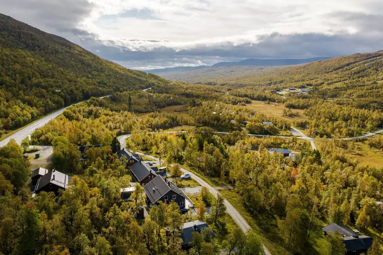 Vacker höstvy över Buskvallen Inn, omgivet av gyllene björkar och slingrande vägar. Molnen skapar dramatik över fjällen.