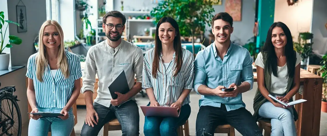 Pięcioro młodych ludzi z uśmiechem na twarzy, przygotowujących się do pracy w branży marketingowej. Każdy z nich ma przy sobie narzędzia pracy: laptop, tablet, telefon, notatnik.